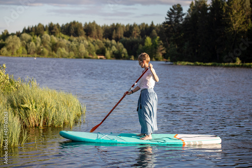 A young girl in a skirt rides a SUP board on a forest pond