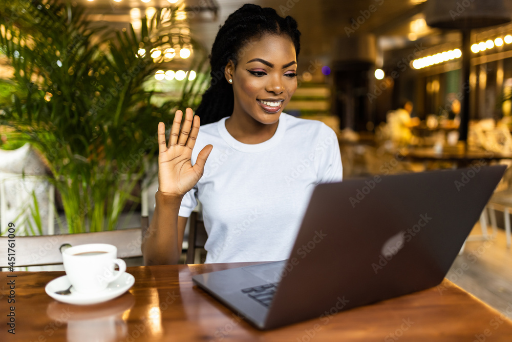 Smiling African woman, young female student with Afro hair talking, waving hand, using laptop computer video conference calling greeting friend by webcam virtual meeting sits at table in cafe. Smiling African woman, young female student with Afro hair talking, waving hand, using laptop computer video conference calling greeting friend by webcam virtual meeting sits at table in cafe.