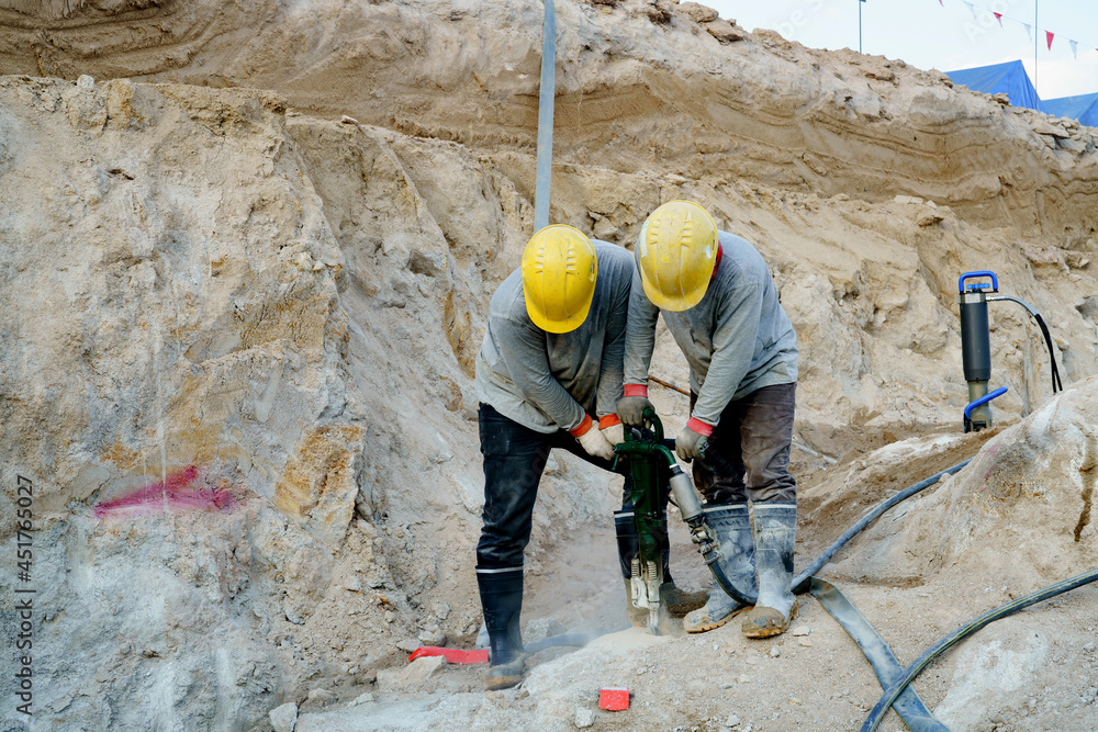 Two builders work together with a large rock drill to break apart in a construction site. Two builders work together with a large rock drill to break apart in a construction site.