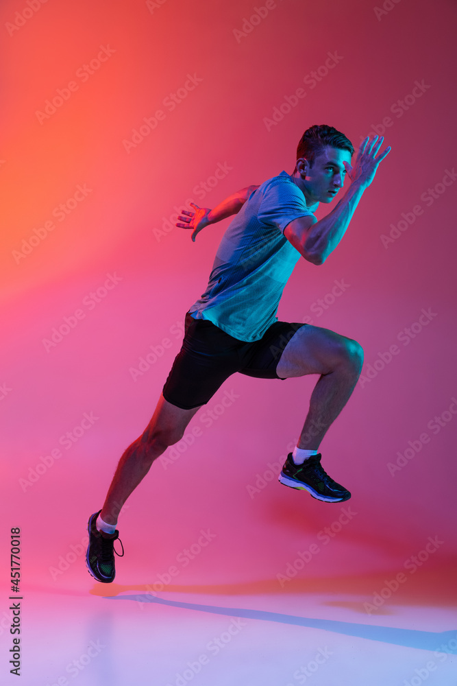 Portrat of Caucasian professional male athlete, runner training isolated on pink studio background with blue neon filter, light. Muscular, sportive man.