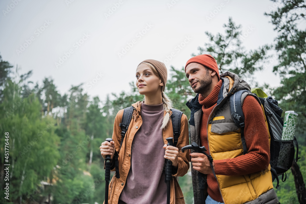 couple with backpacks holding hiking sticks and looking away in forest