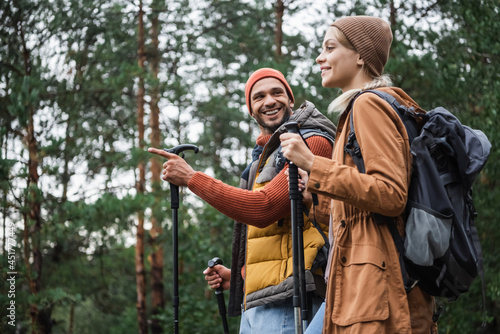 happy man pointing away with finger while looking at girlfriend with hiking sticks in forest