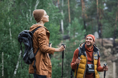 happy man with vintage camera looking at woman holding hiking sticks in forest