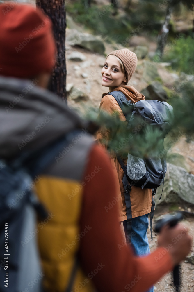 Naklejka premium happy woman with backpack looking at man trekking in forest on blurred foreground