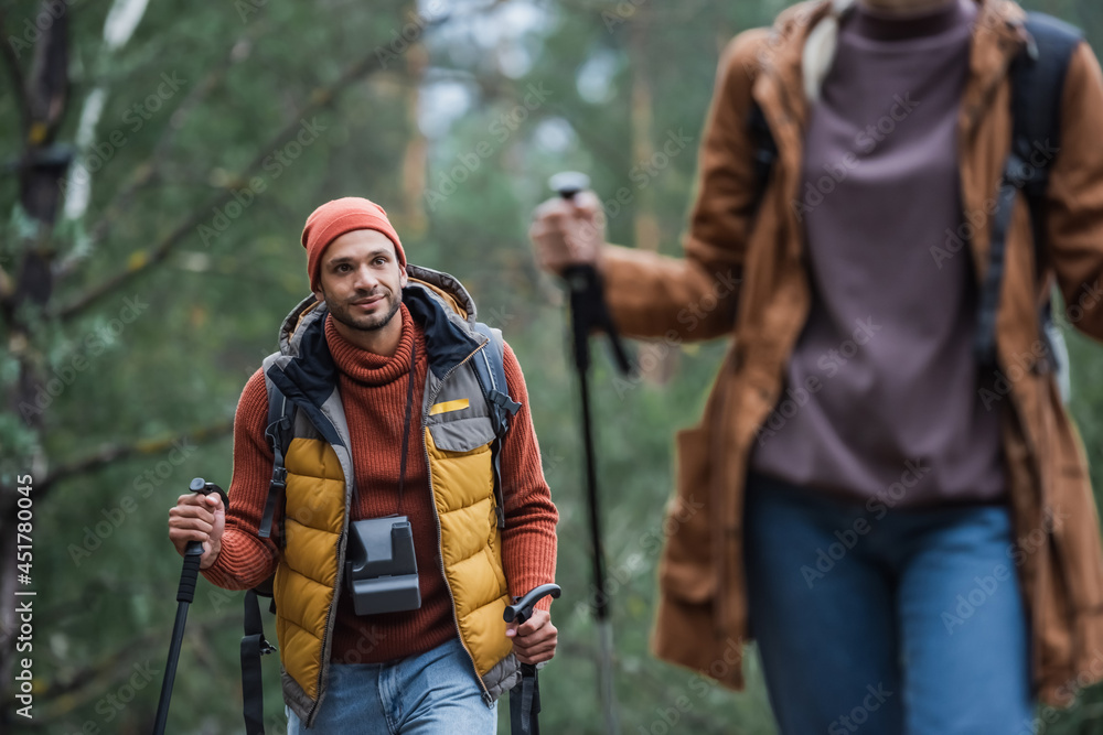 Fototapeta premium man with vintage camera looking at woman holding hiking sticks while trekking in forest on blurred foreground