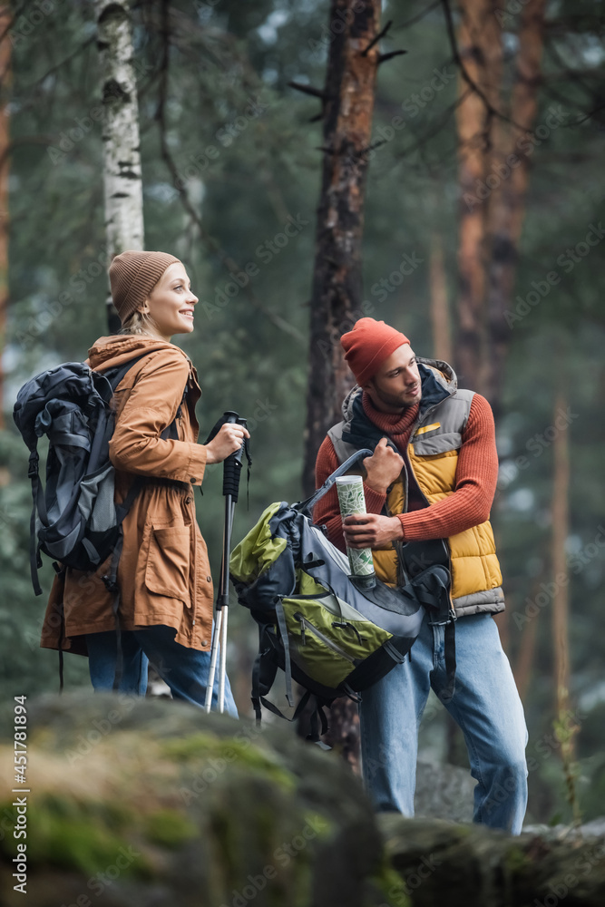 man holding map and backpack near cheerful woman with hiking sticks