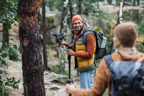 smiling man holding vintage camera and looking at woman trekking in forest on blurred foreground