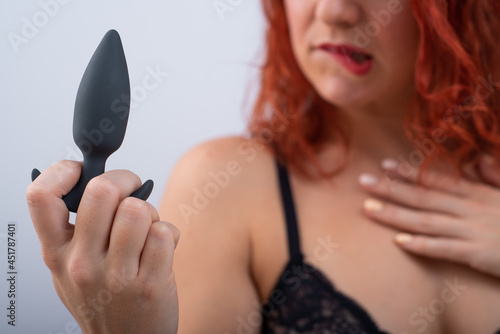 Excited red-haired woman in underwear holds a black anal plug in her hands on a white background