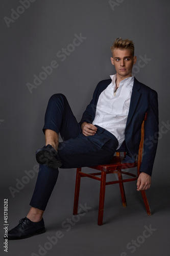 young guy in a blue suit and white shirt posing while sitting in the studio on a gray background © Alexander