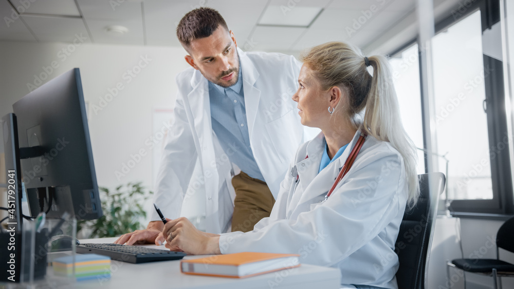 Hospital Medical Doctor's Office: Two Professional Physicians Talking, Solving Problem, Working on Computer. Specialists Discuss Patient Online Treatment, Writing Digital Medicine Prescriptions