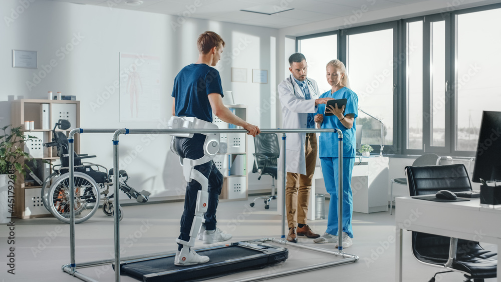 Modern Hospital Physical Therapy: Patient with Injury Walks on Treadmill Wearing Advanced Robotic Exoskeleton. Physiotherapy Rehabilitation Scientists, Engineers, Doctors Help Disabled Person Modern Hospital Physical Therapy: Patient with Injury Walks on Treadmill Wearing Advanced Robotic Exoskeleton. Physiotherapy Rehabilitation Scientists, Engineers, Doctors Help Disabled Person