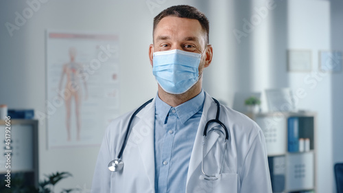 Portrait of Medical Doctor Wearing Face Mask, White Coat and Stethoscope, Standing in His Health Clinic Office. Successful Physician Looks at the Camera. Covid-19, Coronavirus Safety and Protection