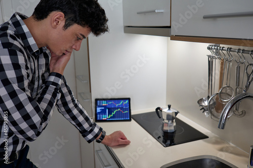 worried young man on phone while making coffee in kitchen and stock data on screen