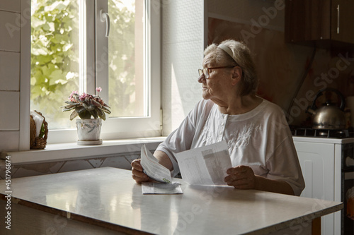 Elderly woman sitting at table in kitchen at home holds domestic bills, feels concerned forgot to pay or debt formed. Middle aged 80s retired woman