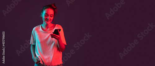 Portrait of pretty romantic girl with phone, gadgets isolated on dark purple studio background in red neon light, filter. Concept of human emotions, facial expression.