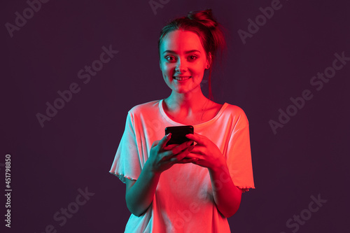 Portrait of pretty romantic girl with phone, gadgets isolated on dark purple studio background in red neon light, filter. Concept of human emotions, facial expression.