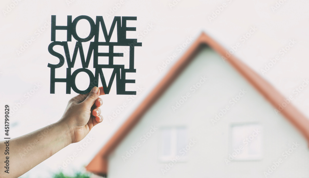 Hand holds wooden phrase Home sweet home in front of blurred house. Concept of buying or building home of dreams Hand holds wooden phrase Home sweet home in front of blurred house. Concept of buying or building home of dreams