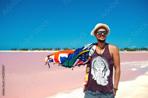 Happy young man in sunglasses and straw hat smiling and posing against pink salt lake in Las Coloradas Mexico, beautiful blue sky 