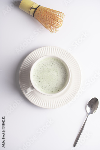 A white porcelain cup with japanese matcha tea drink on a white saucer plate on a white surface, a tea spoon with matcha powder and bamboos whisker , top view