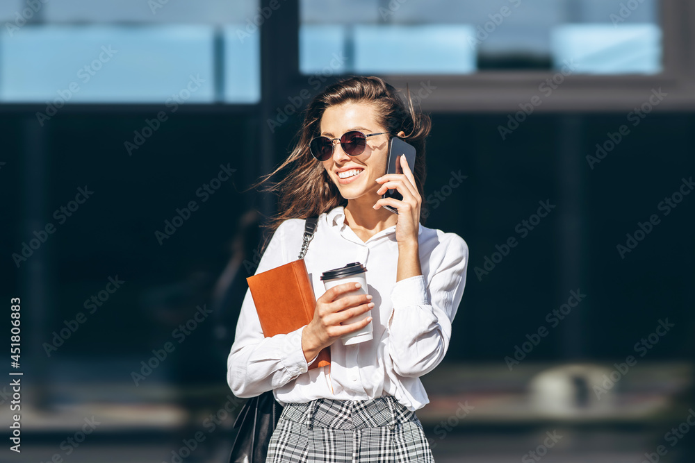 Fototapeta premium Business woman walking and talking on the cellphone outdoors near modern business center with notebook.