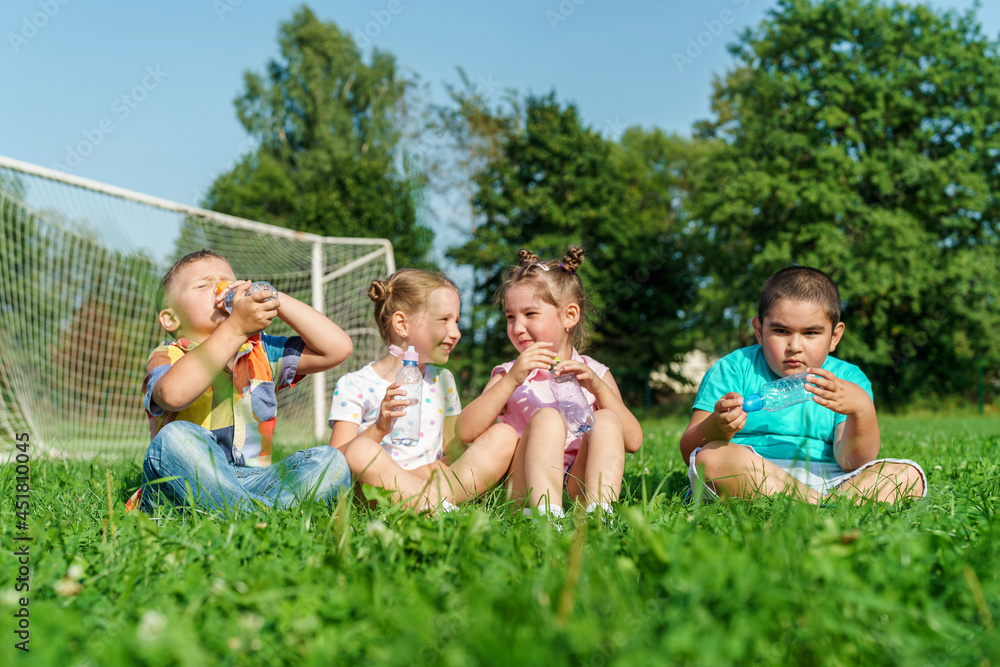 Obraz premium Small children, preschoolers drink water sitting on grass in park on sunny summer day. Outdoor games in fresh air. fun childhood. An active game. Water balance. children are very thirsty.