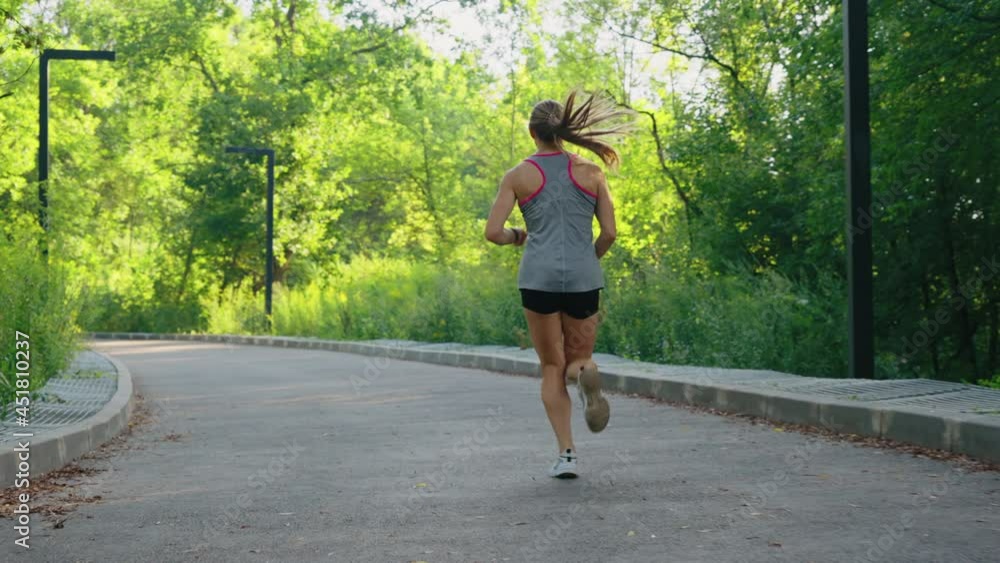 Following shot of blonde woman with ponytail wearing sports shorts and top jogging on rounded road in slow motion. Female athlete from behind training outside on sunny morning. Concept of fitness Following shot of blonde woman with ponytail wearing sports shorts and top jogging on rounded road in slow motion. Female athlete from behind training outside on sunny morning. Concept of fitness