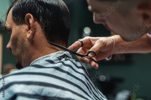 young barber guy gives a haircut to a bearded man sitting in a chair in a barbershop. A man cuts his hair with scissors and a comb. Men's haircuts and beard shaving. Haircut and edging of eyebrows.