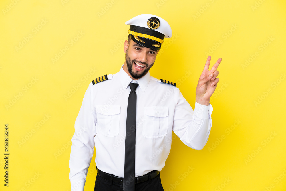 Airplane arab pilot man isolated on yellow background smiling and showing victory sign Airplane arab pilot man isolated on yellow background smiling and showing victory sign