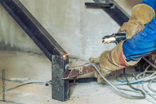 Working male welder with the help of a welding machine assembles the metal structure of the future staircase in a residential building. Sparks of hot metal are flying.