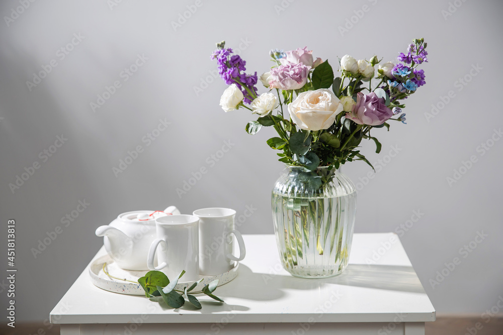 Bouquet of hackelia velutina, purple and white roses, small tea roses, matthiola incana and blue iris in glass vase is on the white coffee table. Grey wall