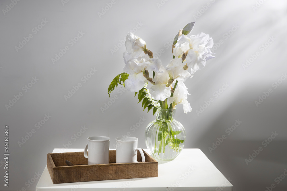 A bouquet of three white irises and a fern in a transparent vase on the table. Two ceramic tea in the tray. Breakfast A bouquet of three white irises and a fern in a transparent vase on the table. Two ceramic tea in the tray. Breakfast