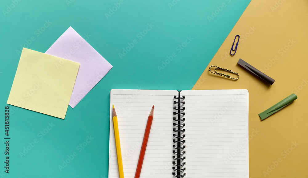 Top view flat lay of blank page notebook in a line with colored pencils, paper clips and pencil caps on green and yellow background Top view flat lay of blank page notebook in a line with colored pencils, paper clips and pencil caps on green and yellow background