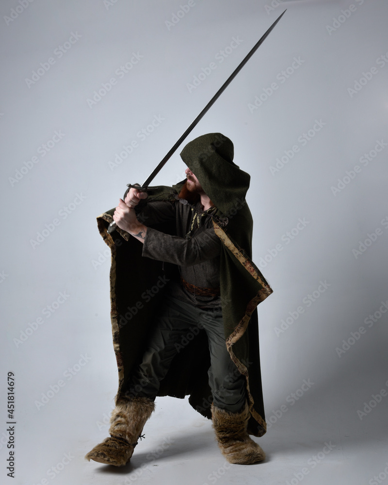 Full length  portrait of  young handsome man  wearing  medieval Celtic adventurer costume with hooded cloak, holding sword, isolated on studio background.