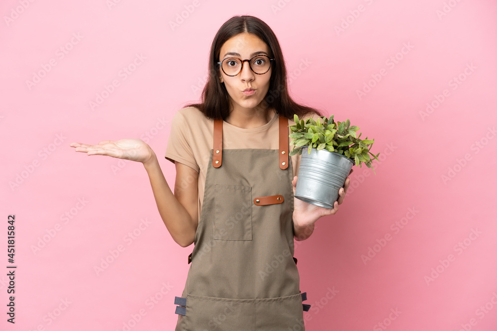 Young gardener girl holding a plant isolated on pink background having doubts while raising hands Young gardener girl holding a plant isolated on pink background having doubts while raising hands