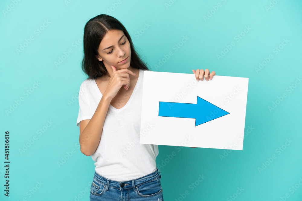 Young French woman isolated on blue background holding a placard with arrow symbol and thinking Young French woman isolated on blue background holding a placard with arrow symbol and thinking