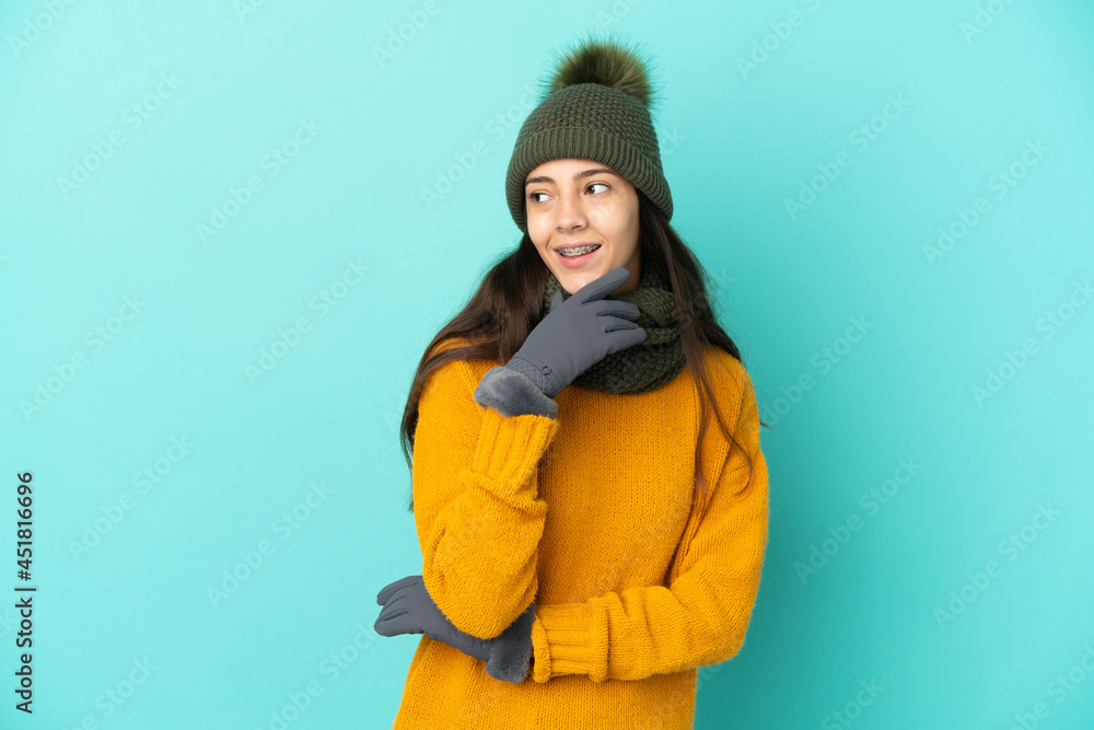 Young French girl isolated on blue background with winter hat looking up while smiling Young French girl isolated on blue background with winter hat looking up while smiling