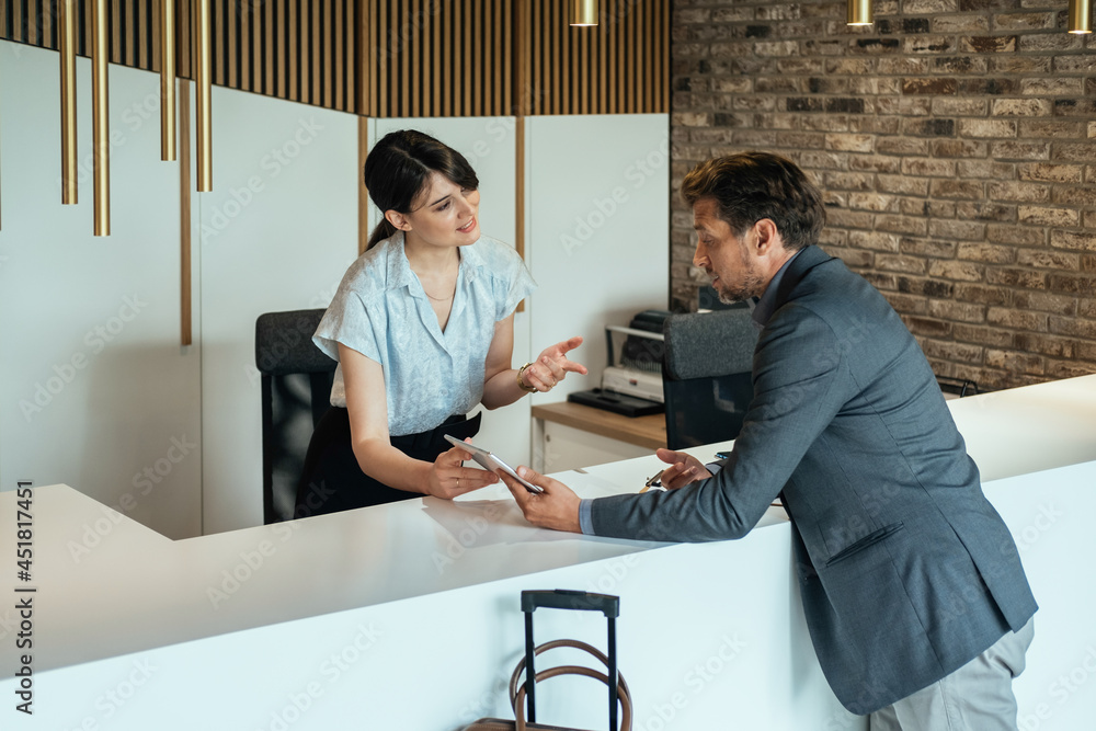Female Hotel Receptionist Assisting Businessman for Checking In at Hotel on Digital Tablet.
Smiling female concierge standing at hotel reception  and showing available rooms on digital tablet to guest - obrazy, fototapety, plakaty 
