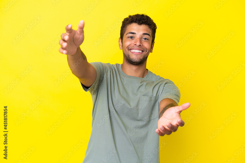 Young handsome Brazilian man isolated on yellow background presenting and inviting to come with hand Young handsome Brazilian man isolated on yellow background presenting and inviting to come with hand