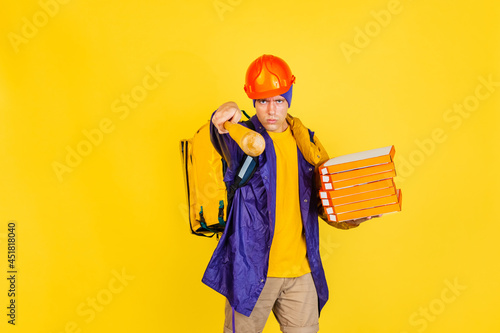 Comic portrait of young man, delivery guy in uniform isolated on yellow studio background. Concept of humor, safety, service. Complementary colors photo