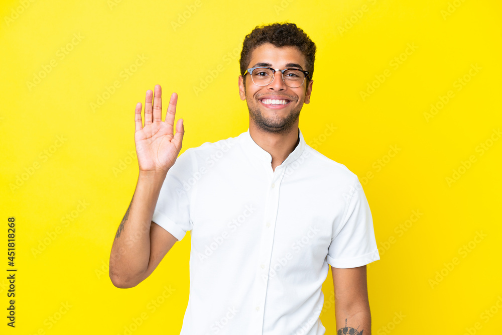 Young handsome Brazilian man isolated on yellow background saluting with hand with happy expression Young handsome Brazilian man isolated on yellow background saluting with hand with happy expression