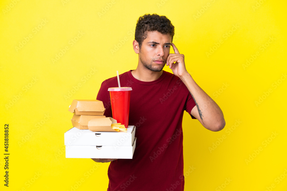 Young brazilian man holding pizzas and burgers isolated background having doubts and thinking Young brazilian man holding pizzas and burgers isolated background having doubts and thinking