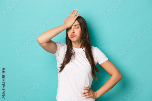 Young tired woman in white t-shirt, sighing and making facepalm to express annoyance, feeling fed up and tensed, standing over blue background photo