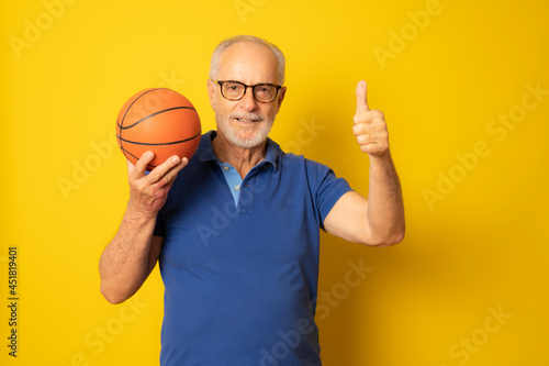 Joyful senior man in casual polo shirt holding a ball and giving a thumb up isolated on yellow background © Danko