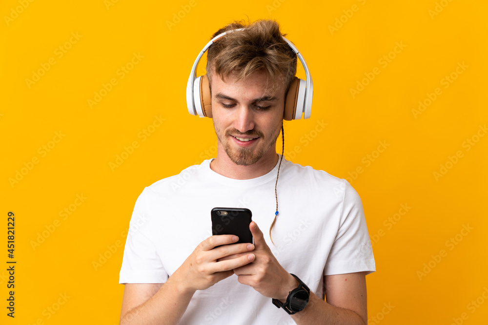 Young handsome blonde man isolated on yellow background listening music and looking to mobile
