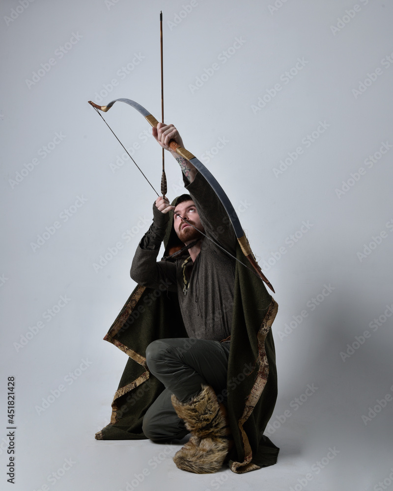 Full length portrait of young handsome man wearing medieval Celtic adventurer costume with hooded cloak, holding a archery bow and arrow, isolated on studio background. Full length portrait of young handsome man wearing medieval Celtic adventurer costume with hooded cloak, holding a archery bow and arrow, isolated on studio background.