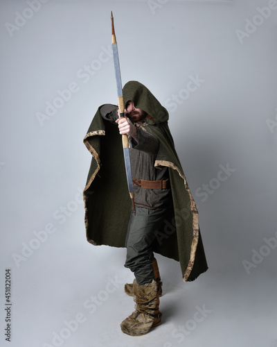 Full length portrait of young handsome man wearing medieval Celtic adventurer costume with hooded cloak, holding a archery bow and arrow, isolated on studio background.