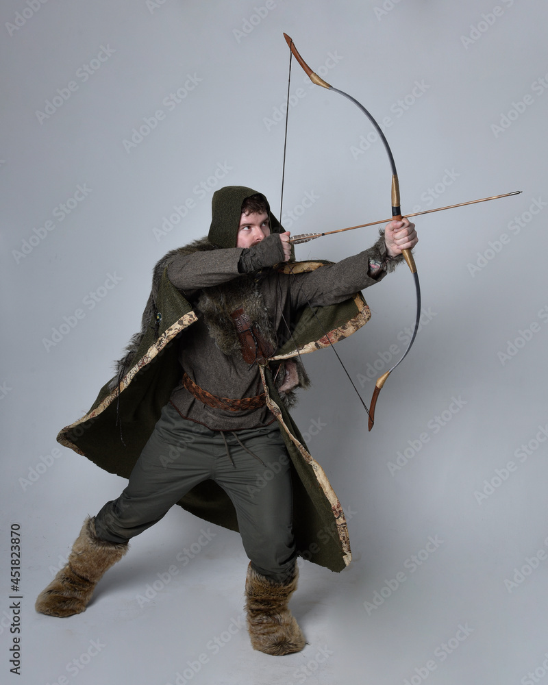 Full length portrait of young handsome man wearing medieval Celtic adventurer costume with hooded cloak, holding a archery bow and arrow, isolated on studio background. Full length portrait of young handsome man wearing medieval Celtic adventurer costume with hooded cloak, holding a archery bow and arrow, isolated on studio background.