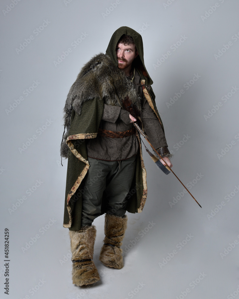 Full length portrait of young handsome man wearing medieval Celtic adventurer costume with hooded cloak, holding a archery bow and arrow, isolated on studio background. Full length portrait of young handsome man wearing medieval Celtic adventurer costume with hooded cloak, holding a archery bow and arrow, isolated on studio background.