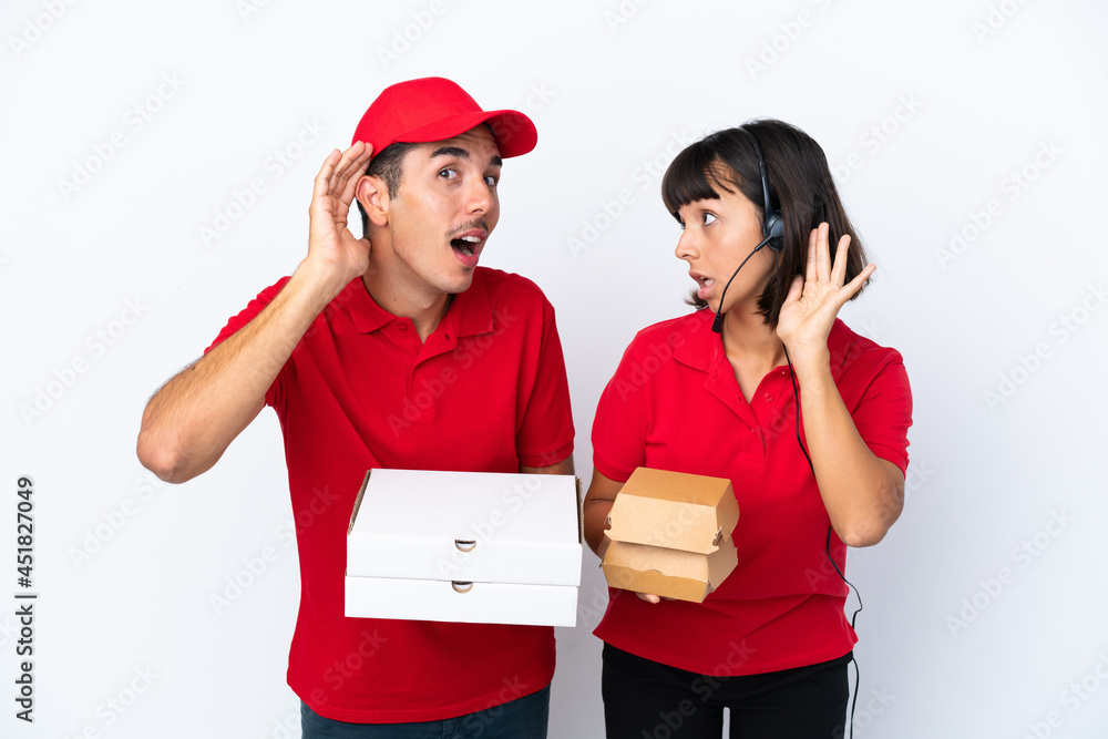 Young delivery couple holding pizzas and burgers isolated on white background listening to something by putting hand on the ear Young delivery couple holding pizzas and burgers isolated on white background listening to something by putting hand on the ear