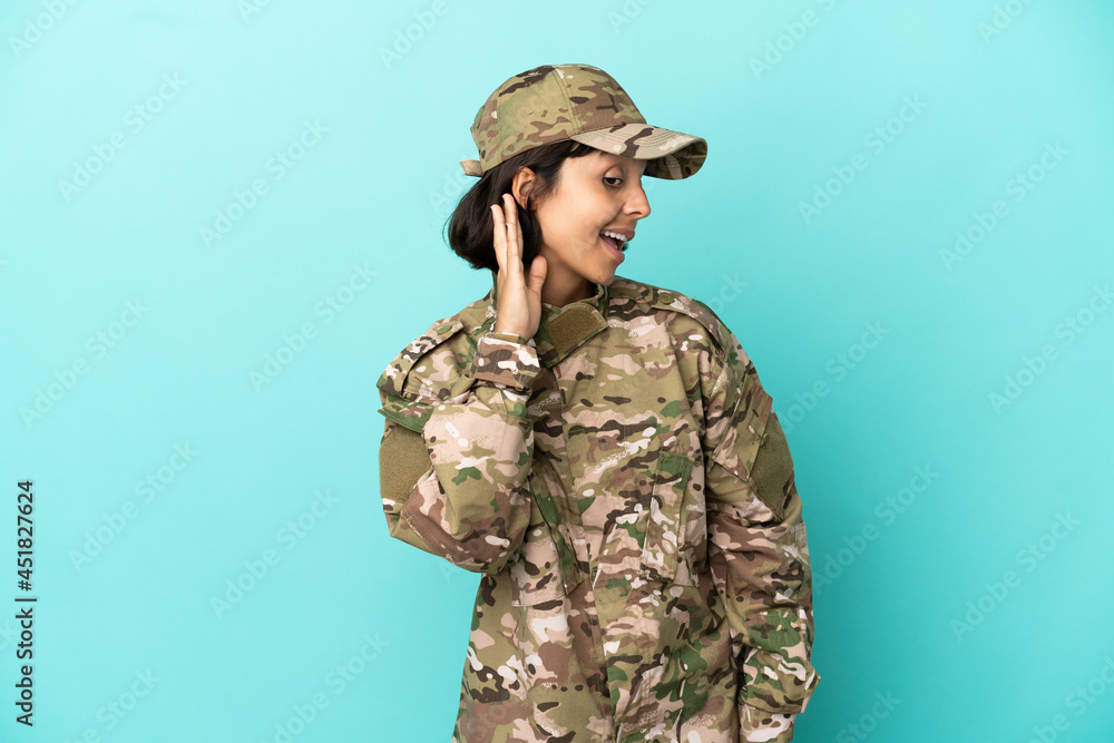 Military mixed race woman isolated on blue background listening to something by putting hand on the ear